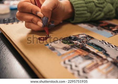 Close-up image of an unrecognizable young woman's hand filling in a red location mark with a felt-tip pen on a handmade kraft album with travel photos and washi tape. Contains the word market in Thai.