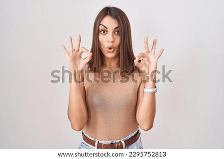 Young hispanic woman standing over white background looking surprised and shocked doing ok approval symbol with fingers. crazy expression 