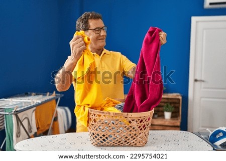 Middle age man smiling confident holding clothes of basket at laundry room