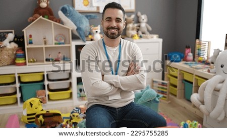 Young hispanic man teacher smiling confident sitting with arms crossed gesture at kindergarten