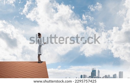 Confident medical industry employee in white suit standing at the top of building roof and holding tablet in hands. Skyscape and city view on background. Medical sphere concept Royalty-Free Stock Photo #2295757425
