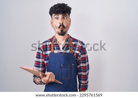 Young hispanic man with beard wearing waiter apron holding clipboard making fish face with lips, crazy and comical gesture. funny expression.  Royalty-Free Stock Photo #2295761569