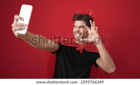Young hispanic man wearing devil costume make selfie by smartphone over isolated red background