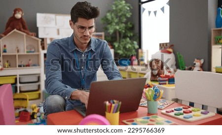 Young hispanic man preschool teacher using laptop sitting on table at kindergarten Royalty-Free Stock Photo #2295766369