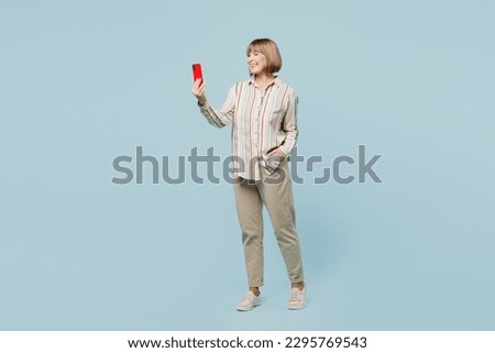 Full body fun elderly woman 50s years old wear shirt hold in hand use mobile cell phone doing selfie shot isolated on plain pastel light blue cyan color background studio portrait. Lifestyle concept