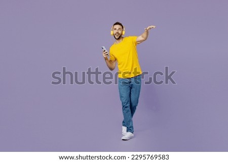 Full body young smiling happy fun man wearing yellow t-shirt headphones listening music use mobile cell phone dance isolated on plain pastel light purple background studio portrait. Lifestyle concept