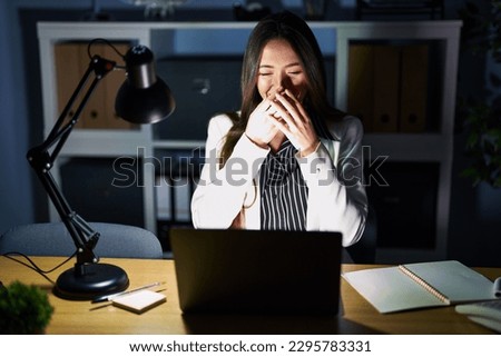Young brunette woman working at the office at night with laptop laughing and embarrassed giggle covering mouth with hands, gossip and scandal concept 