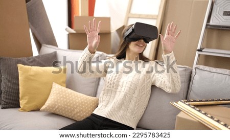Young beautiful hispanic woman using virtual reality glasses sitting on sofa at new home