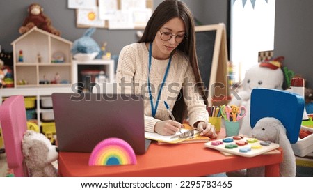 Young beautiful hispanic woman preschool teacher using laptop writing on document at kindergarten