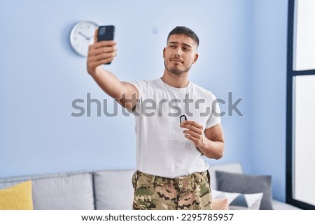 Young hispanic man wearing camouflage army uniform taking selfie at home smiling looking to the side and staring away thinking. 