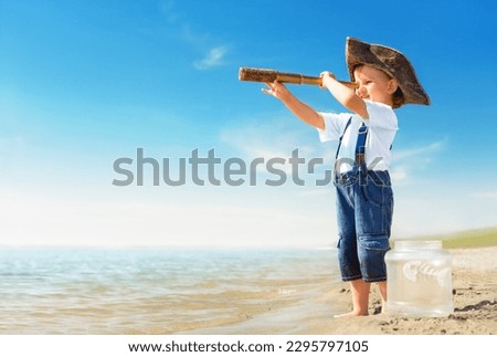 Kid play on the beach on a hot sunny day. Little girl dressed as a pirate stands barefoot on the sandy seashore with a telescope. Child with a toy fish dreams of travel and adventure.