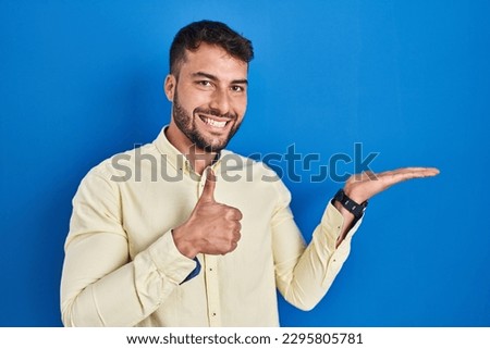 Handsome hispanic man standing over blue background showing palm hand and doing ok gesture with thumbs up, smiling happy and cheerful 
