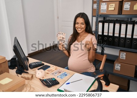 Young pregnant woman working at small business ecommerce holding mexican pesos smiling happy and positive, thumb up doing excellent and approval sign 