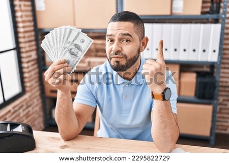 Hispanic man working at small business ecommerce holding dollars smiling with an idea or question pointing finger with happy face, number one 