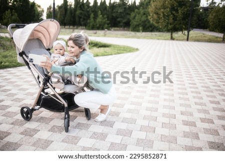 Young mother taking a selfie with her infant. The baby sits in a stroller. Mom is on maternity leave. Stylish mom in a knitted sweater and white trousers, baby in a pink eco knitted combination