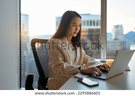 Busy Asian business woman using laptop in company office. Young female digital finance professional worker using computer doing corporate analysis online management sitting at desk, city window view. Royalty-Free Stock Photo #2295886601