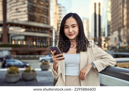Young smiling pretty Asian business woman professional office manager outdoors with cellphone in hand standing on big city street holding mobile cell phone device, using smartphone, portrait.