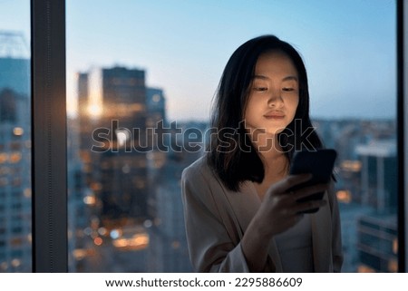 Young busy Asian business woman executive using mobile cell phone technology at night in dark office. Professional lady businesswoman working on digital smartphone with evening city view from window. Royalty-Free Stock Photo #2295886609