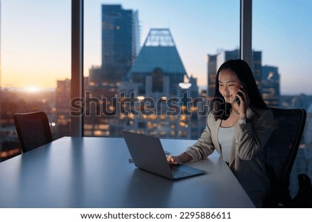Young busy Asian business woman executive working on laptop making call at night in dark corporate office. Professional businesswoman manager talking to client using computer, city evening window view Royalty-Free Stock Photo #2295886611