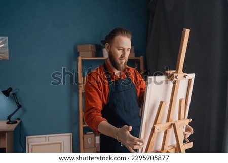 portrait caucasian bearded male artist in an apron in a home studio stands near an easel holds a canvas in his hands and looks at the picture contemptuously. hobby and vacation concept. Royalty-Free Stock Photo #2295905877
