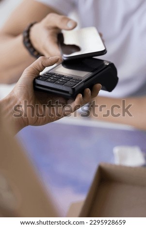Close up hand of male customer makes transaction, holds mobile phone applied modern gadget on payment terminal equipment, paying for services using NFC technology, easy contactless cashless e-payment