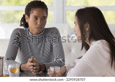 Serious African woman spend time with girlfriend seated at table with beverages in cozy cafe, share problems, ask for advice, discuss personal life concerns to best reliable friend, having honest talk Royalty-Free Stock Photo #2295928707