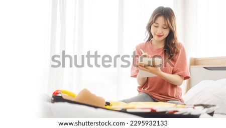 Portrait of young adult southeast asian dyed hair woman with travel luggage. Thinking about backpack with digital tablet for trip check list. Sitting on bed at home or hotel bedroom. Royalty-Free Stock Photo #2295932133