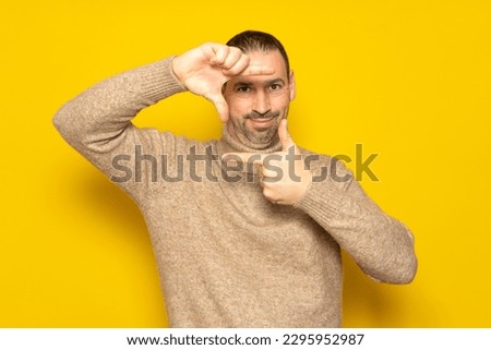 Handsome bearded happy male blink with eye and looking through a frame formed by his hands. Attractive man make frame by fingers, isolated on yellow background with copy space for text