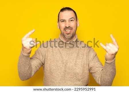 Photo of excited funky guy dressed in beige turtleneck sweater showing two heavy rock gestures over isolated yellow color background.