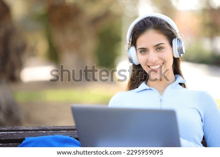 Happy student with laptop and headphone looks at you in a park