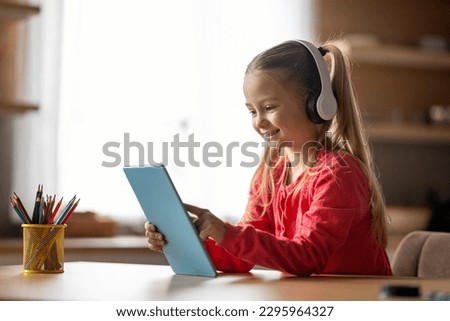Cute Little Girl In Wireless Headphones Relaxing With Digital Tablet At Home, Cheerful Preteen Female Child Playing Games Or Browsing Internet On Modern Gadget, Sitting At Desk Indoors, Copy Space