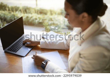 Serious young black woman in white suit at table with computer with empty screen make notes, planning startup in cafe with plants interior, blurred. Device for business and work, create idea