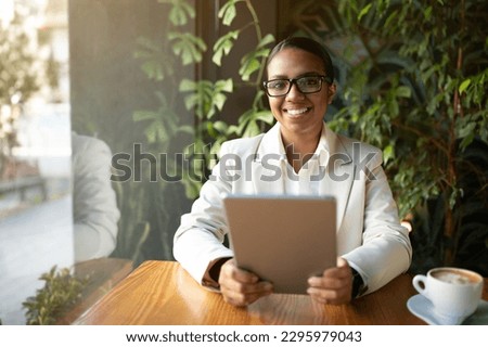 Positive young black lady manager in white suit and glasses sits at table with tablet and cup of coffee in cafe with plants interior. Restaurant blog, work and business, study remotely and app Royalty-Free Stock Photo #2295979043