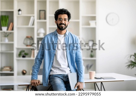 Modern Freelancer. Young Indian Man Standing Near Desk At Home Office, Handsome Millennial Eastern Male Holding Laptop In Hand And Smiling At Camera, Enjoying Distant Work Opportunities, Copy Space Royalty-Free Stock Photo #2295979055