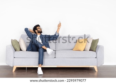 Relaxed handsome bearded middle eastern young man in casual outfit and eyeglasses sitting on couch at home, holding remote control, turning on ac, pointing towards copy space on white wall Royalty-Free Stock Photo #2295979077