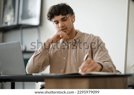 E-Learning. Middle Eastern Man Learning Online Taking Notes Sitting At Laptop Wearing Earbuds At Home. Guy Studying Remotely Using Computer Posing At Desk Indoors. Distance Education Royalty-Free Stock Photo #2295979099