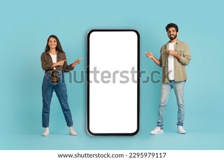 Happy cute beautiful young indian couple in casual pretty woman and handsome guy pointing at big cell phone with white blank screen, mockup for dating app, blue background, full length Royalty-Free Stock Photo #2295979117