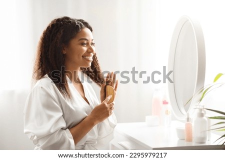 Black Smiling Female Combing Her Beautiful Curly Hair With Bamboo Brush At Home, Attractive African American Woman Sitting At Dressing Table And Looking In Mirror, Making Daily Beauty Routine Royalty-Free Stock Photo #2295979217