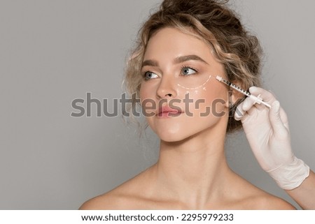 Beautician doctor making injection to young beautiful woman with drawn line under eye, lady with perfect smooth skin getting mesotherapy treatment or hyaluronic acid shot Royalty-Free Stock Photo #2295979233