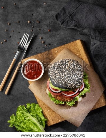 Healthy vegetarian burger with black bun, chickpeas cutlet, salad, cucumber on a cutting board with tomato sauce and spices on dark background. The concept of vegan fast food. Top view.