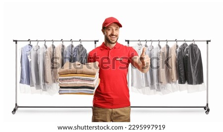 Laundry worker holding a pile of folded clothes and gesturing thumbs up in front of clothing racks isolated on a white background

