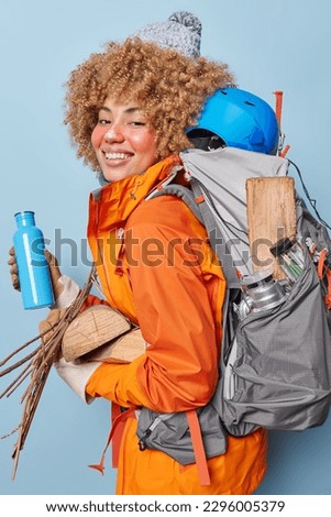 People free time and camping concept. Cheerful female tourist has hiking adventure going to make campfire and have picnic carries wood poses with rucksack full of equipment wears hat and jacket