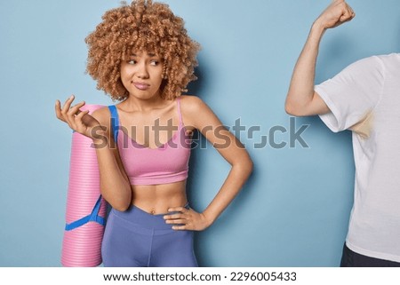 Confused clueless curly haired sportswoman shrugs shoulders with bewilderment looks at someone showing biceps doesnt know which exercise to do first poses with karemat dressed in sportswear. Royalty-Free Stock Photo #2296005433