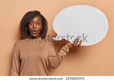 Photo of stunned dark skinned woman with bushy hair has omg expression holds empty speech bubble reacts to shocking news dressed in casual jumper isolated over brown background. Announcement concept
