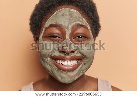 Close up shot of cheerful woman with plump face dark curly hair applies beauty clay mask for rejuvenation has perfect white teeth isolated over brown studio background. Facial cleaning concept