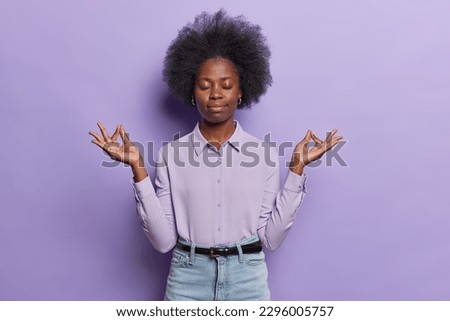 Photo of dark skinned female office worker tries to relax after work makes zen mudra sign inhales positive vibes keeps eyes closed forgets about all troubles wears shirt jeans isolated on purple wall Royalty-Free Stock Photo #2296005757