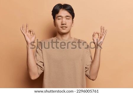 Relieved young Asian man releases stress meditates indoors makes mudra or zen gesture wears casual t shirt stands with closed eyes isolated over brown background practices meditation in studio