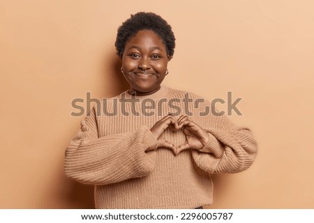 Dark skinned chubby woman with curly short hair makes heart sign gesture expresses love and kindness shows her true feelings looks happily wears knnitted jumper isolated over brown background