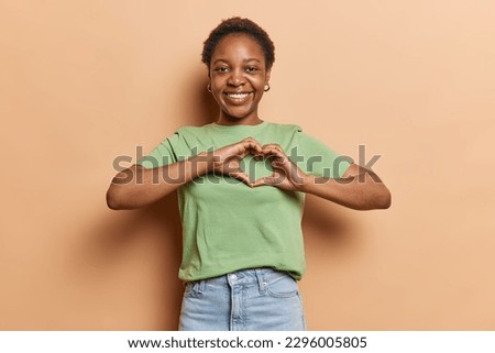 So much love to give. Glad Afro American woman with short hair makes heart shape with her hands has romantic feelings expresses love to you wears casual t shirt and jeans poses against brown wall