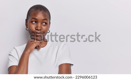 Horizontal shot of pensive dark skinned girl keeps hand under chin concentrared aside wears casual t shirt isolated on white background copy space for your promotional text tries to remember something